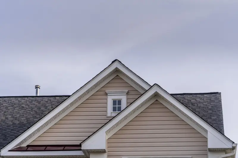 Roofing & Siding Beige horizontal vinyl siding, white decorative frame attic window reverse double gable close up on luxury single family residential home. Square single pane gable window. Roof line of traditional home with one gable running one way reverse gables running the other.